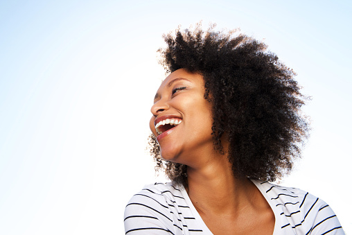 Close up portrait of cheerful young black woman laughing outdoors against bright sky