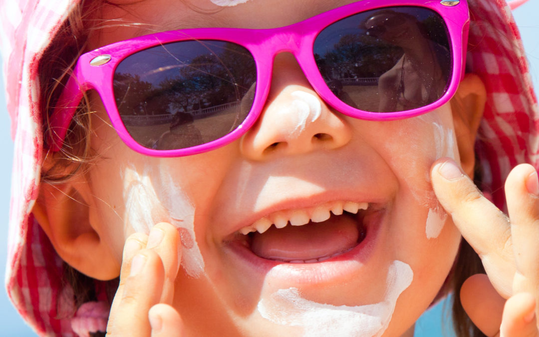 A toddler wearing a hat and sunglasses applying sunscreen to her cheeks.