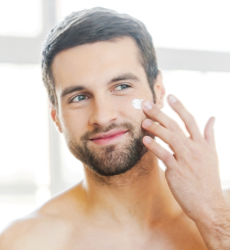 Skin care. Handsome young shirtless man applying cream at his face and looking at himself with smile while standing in front of the mirror